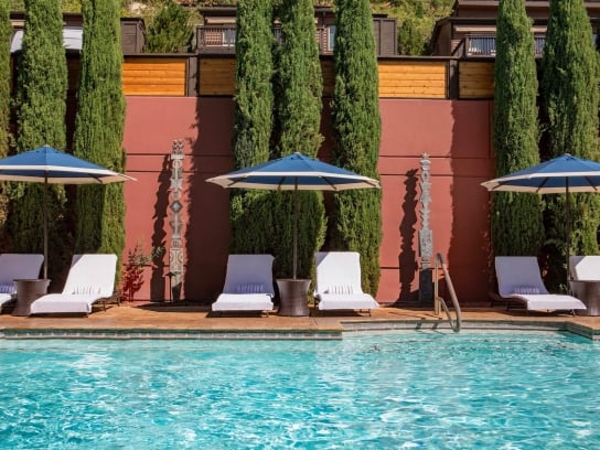 White lounge chairs and blue-striped umbrellas line a sparkling blue pool, set against a red wall and tall cypress trees.