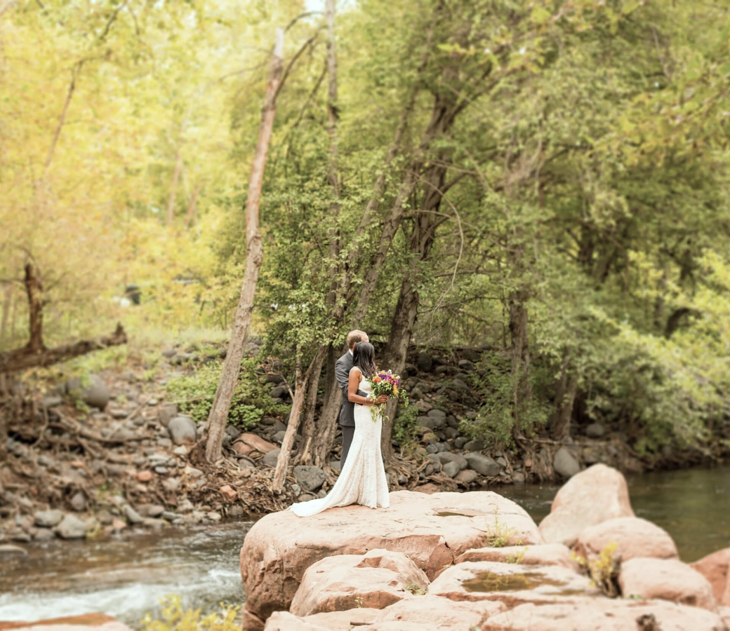 A bride and groom embrace on a large rock in the middle of Oak Creek, surrounded by lush green trees.