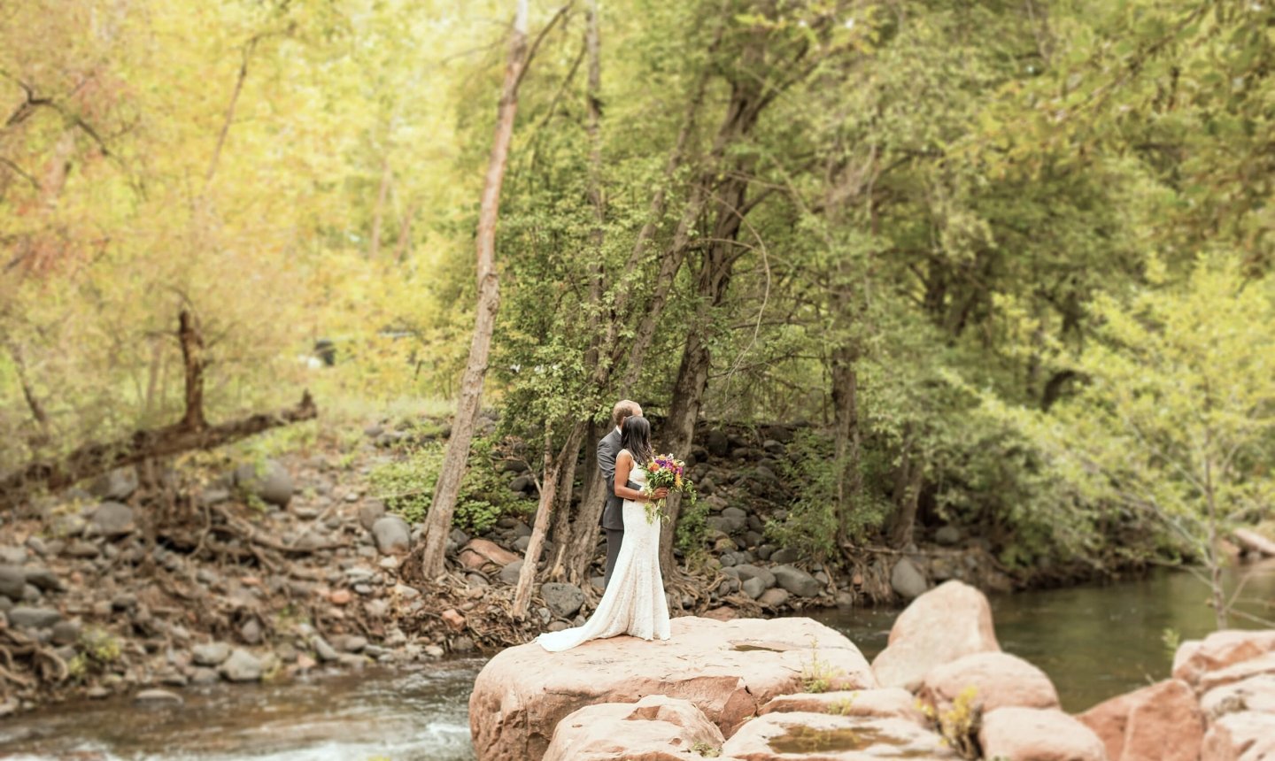 A bride and groom embrace on a large rock in the middle of Oak Creek, surrounded by lush green trees.