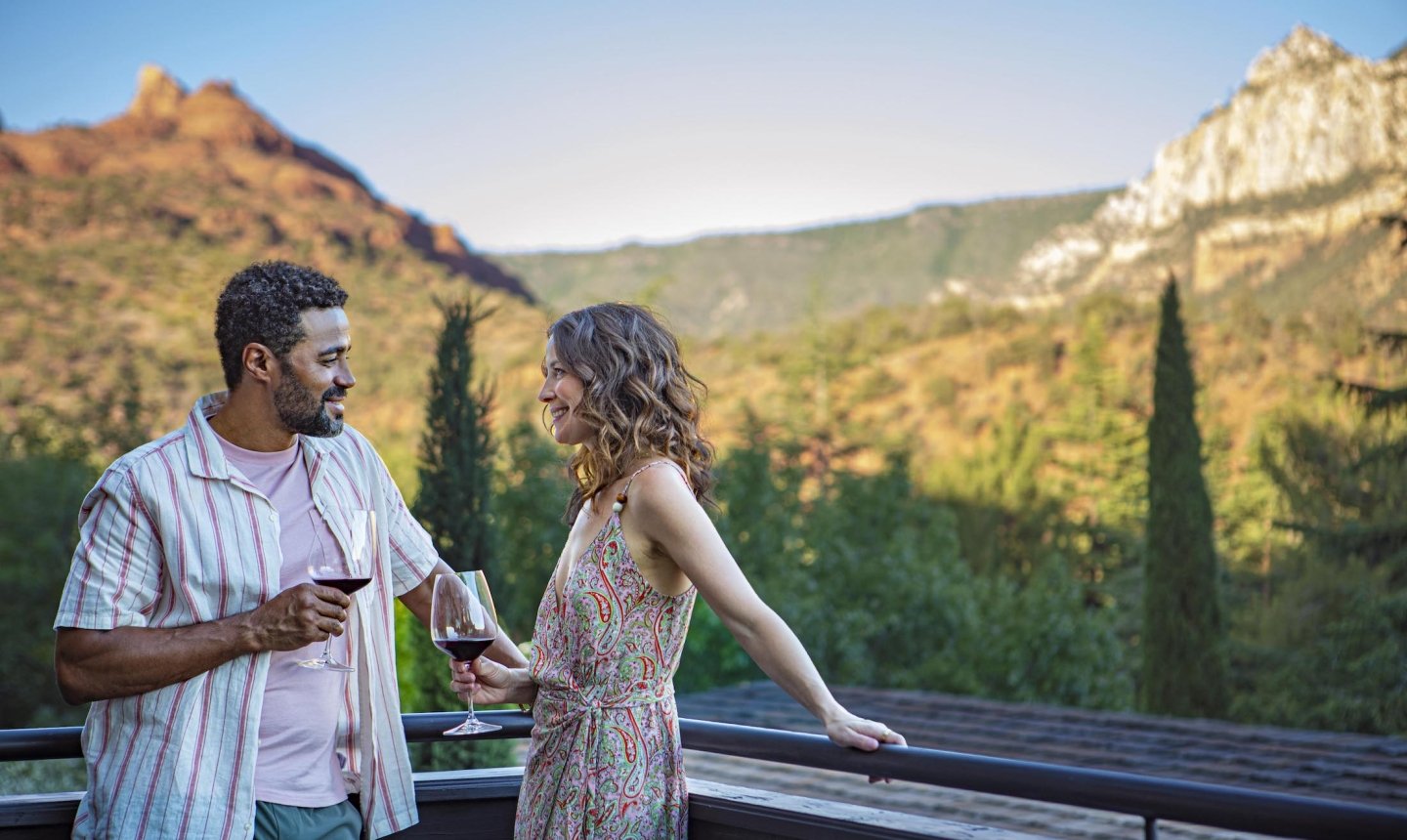 A smiling couple enjoys red wine on a balcony, talking with a panoramic view of Sedona's mountains in the background.