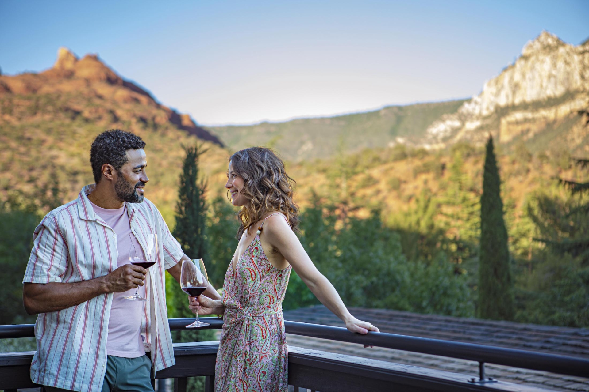 A smiling couple enjoys red wine on a balcony, talking with a panoramic view of Sedona's mountains in the background.
