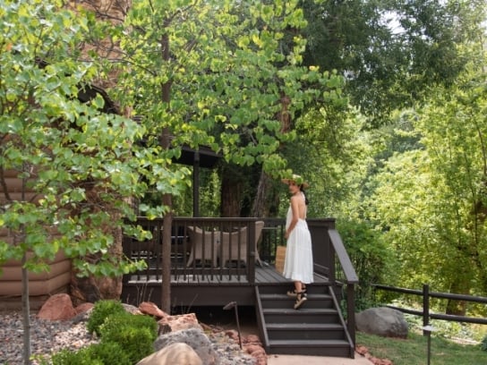 A woman in a white dress and sun hat looks back while walking up the steps to a wooden cottage deck surrounded by trees.