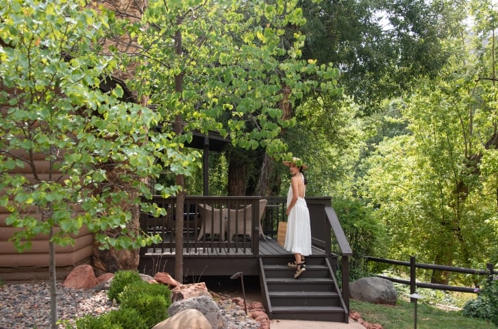 A woman in a white dress and sun hat looks back while walking up the steps to a wooden cottage deck surrounded by trees.