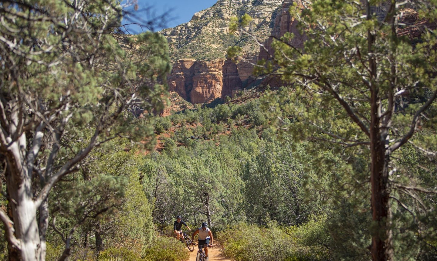 Two people mountain biking on a dirt trail through a green forest, with a large Sedona red rock cliff in the background.