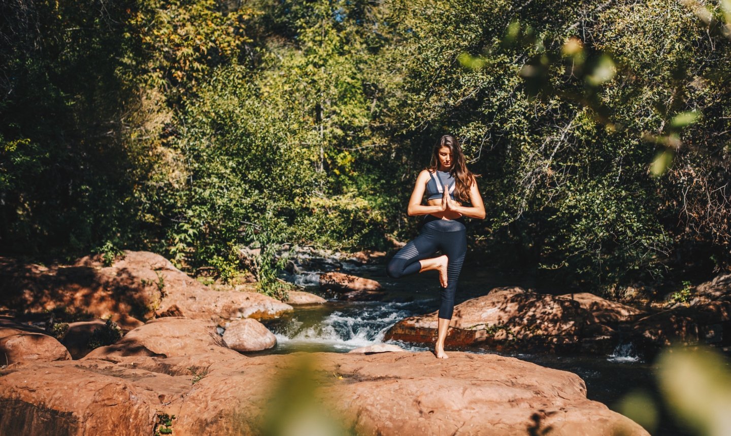 A woman in yoga attire holds a tree pose on a large red rock next to the flowing Oak Creek, surrounded by dense greenery.