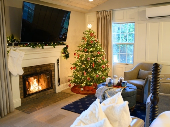 A cozy cottage room decorated for Christmas, with a lit fireplace, white stockings, and a festive, lighted tree.