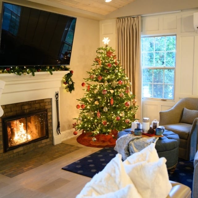 A cozy cottage room decorated for Christmas, with a lit fireplace, white stockings, and a festive, lighted tree.