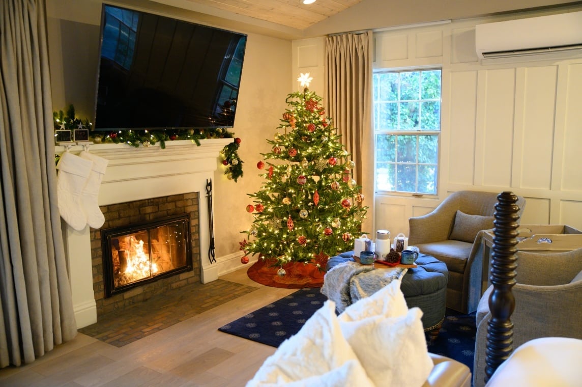 A cozy cottage room decorated for Christmas, with a lit fireplace, white stockings, and a festive, lighted tree.
