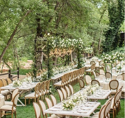 An elegant outdoor wedding reception on a lush green lawn, with long tables and floral arches set among tall trees.