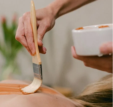 A close-up of a spa treatment, with a therapist applying a body mask to a person's back with a wide brush.