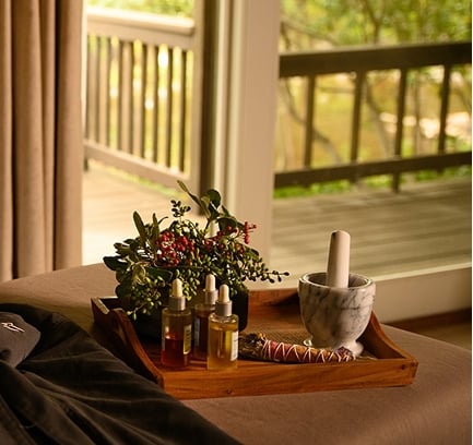 A wooden tray with spa oils, a mortar and pestle, and a smudge stick rests on a table next to a robe, near a balcony.
