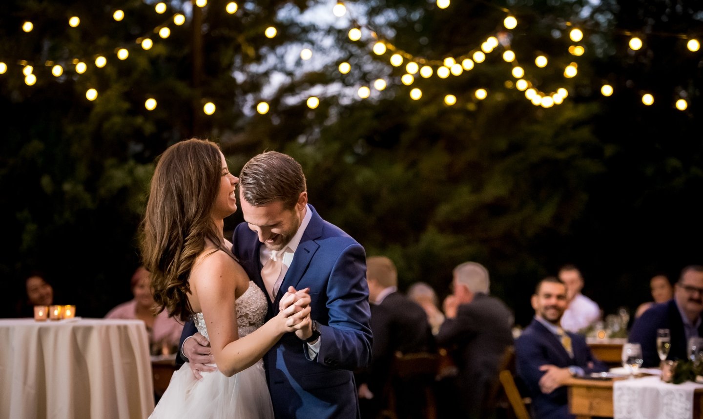 A bride and groom smile and embrace during their first dance at an outdoor wedding reception under string lights.