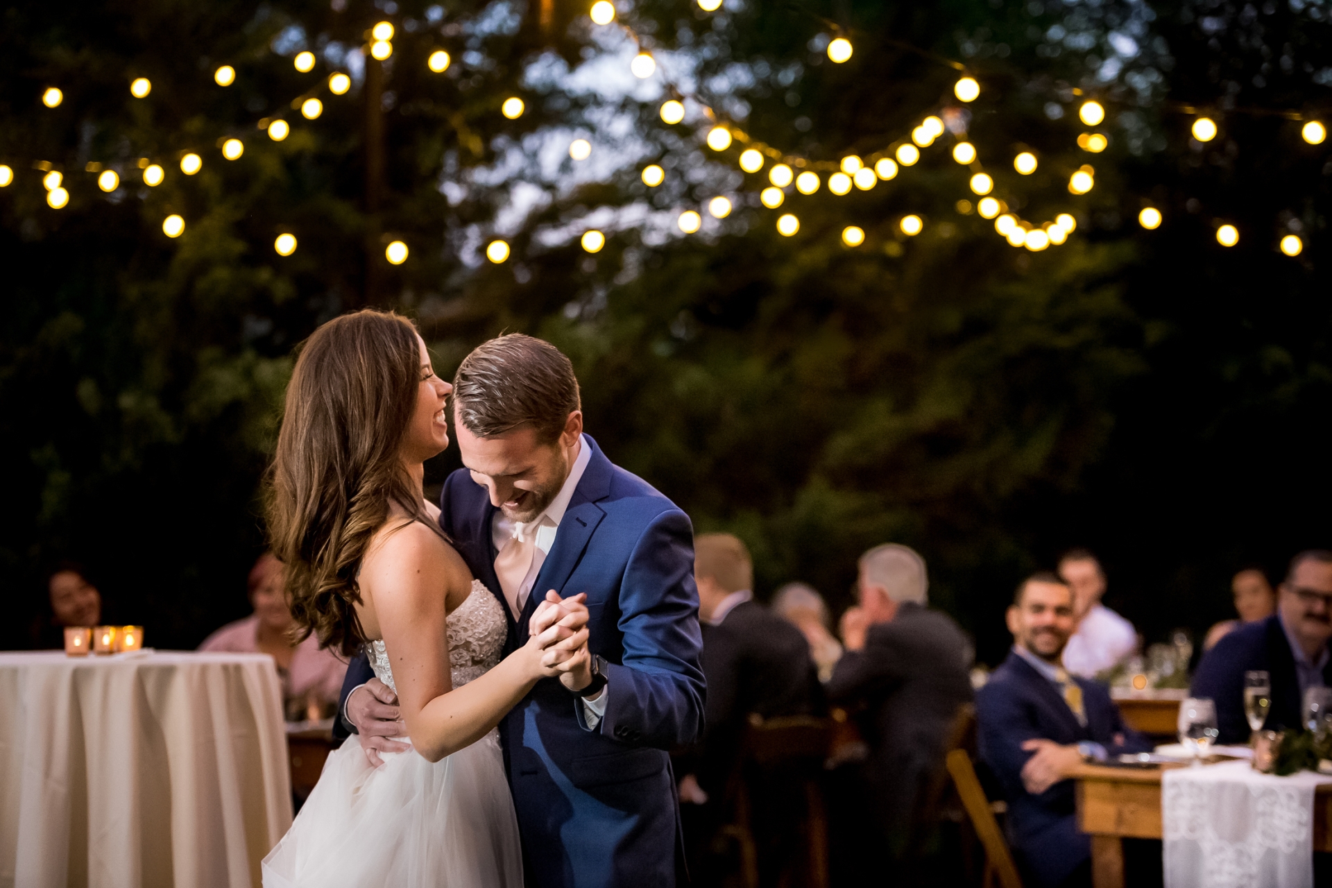 A bride and groom smile and embrace during their first dance at an outdoor wedding reception under string lights.