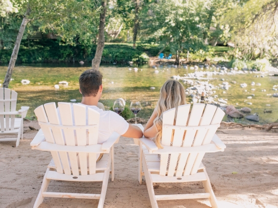 A couple, seen from behind, sits in white Adirondack chairs on a sandy beach, drinking wine by Oak Creek.