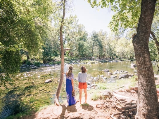 Two women stand on the bank of the rocky and shallow Oak Creek, looking at the water in a sunlit, wooded setting.