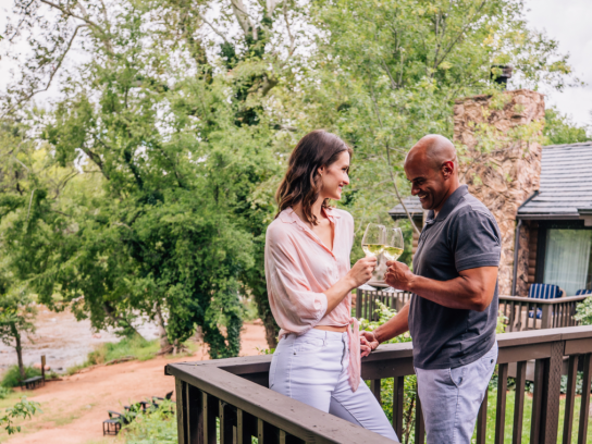 A smiling couple clinks glasses of white wine on a cottage deck, with the lush greenery of Oak Creek behind them.