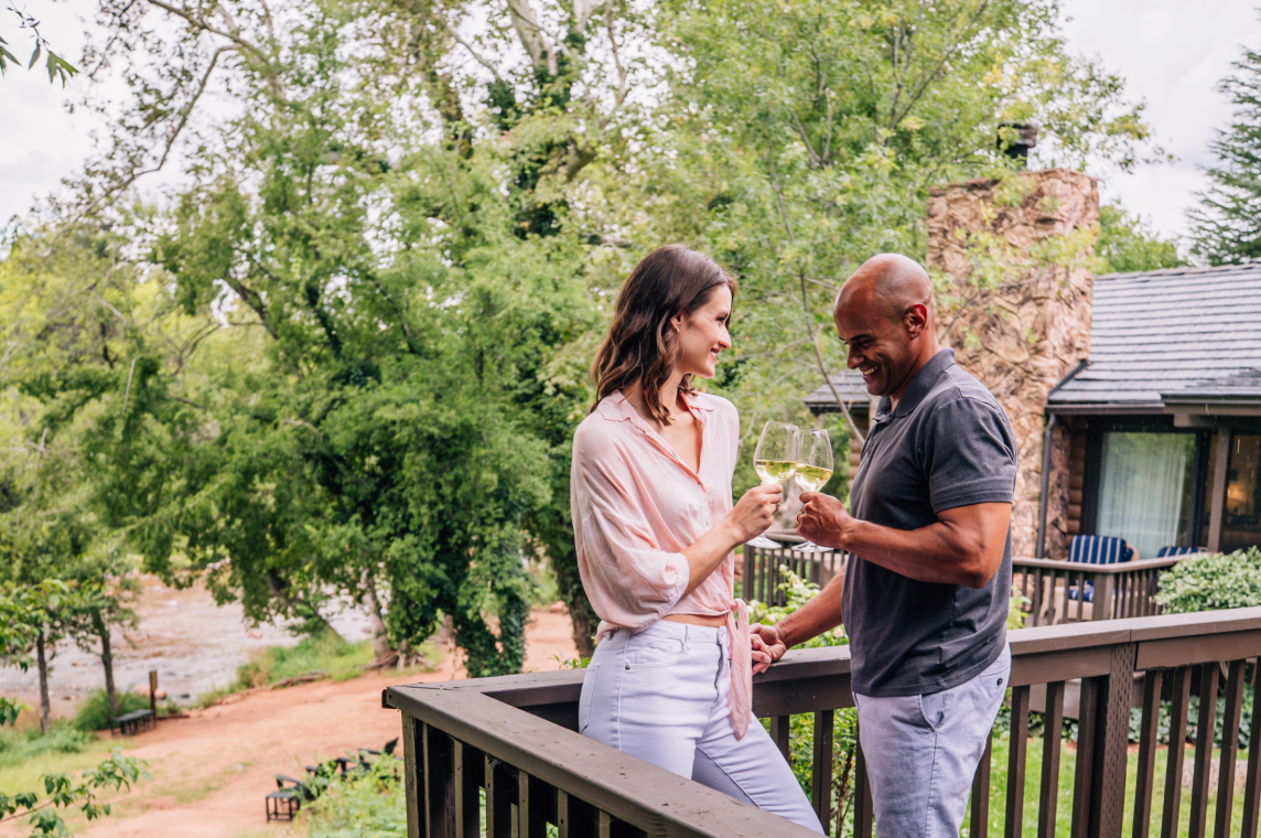 A smiling couple clinks glasses of white wine on a cottage deck, with the lush greenery of Oak Creek behind them.