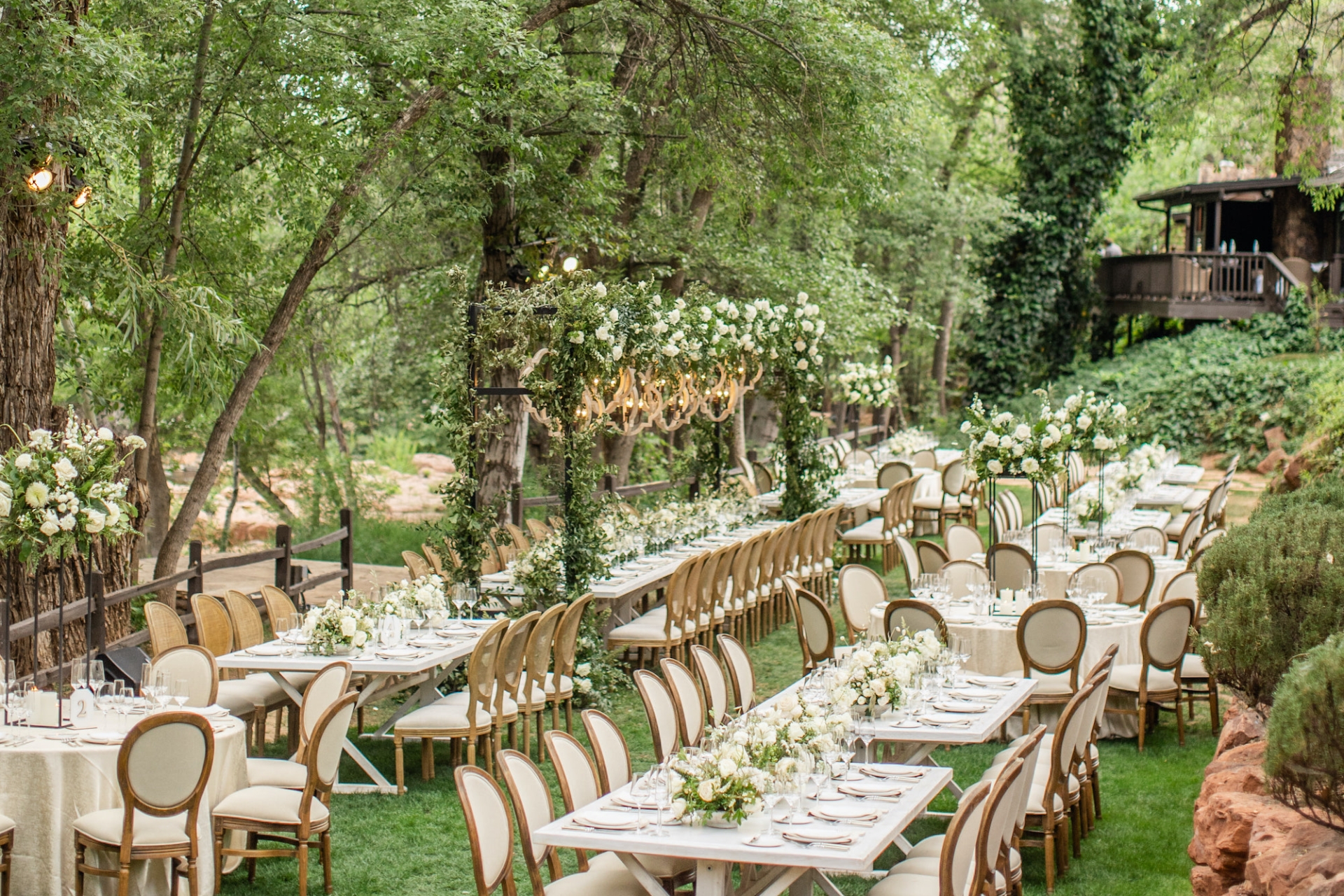 Elegant outdoor wedding reception with long white tables and cushioned chairs, set on a lush green lawn among tall trees.
