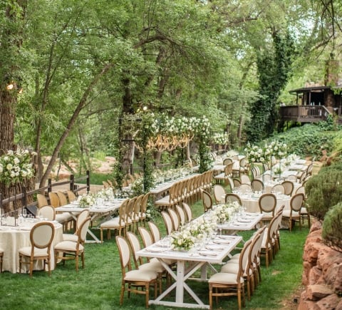Elegant outdoor wedding reception with long white tables and cushioned chairs, set on a lush green lawn among tall trees.