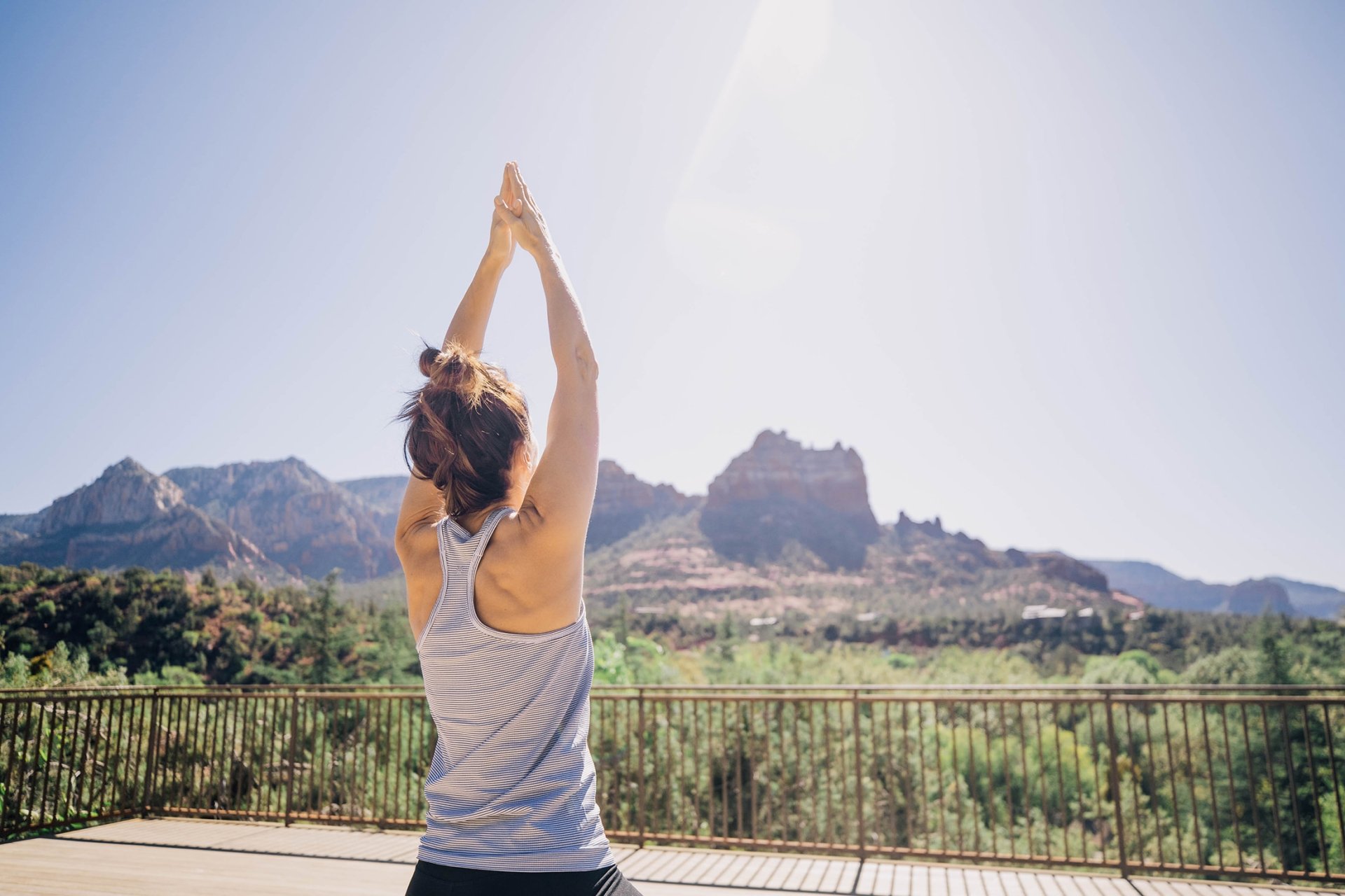 A woman, seen from behind, does a yoga pose with arms raised on a wooden deck overlooking a panoramic view of Sedona's red rocks.