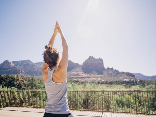A woman, seen from behind, does a yoga pose with arms raised on a wooden deck overlooking a panoramic view of Sedona's red rocks.