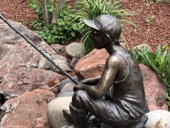 Bronze statue of a barefoot boy fishing, sitting on red rocks by a garden pond with lush greenery.