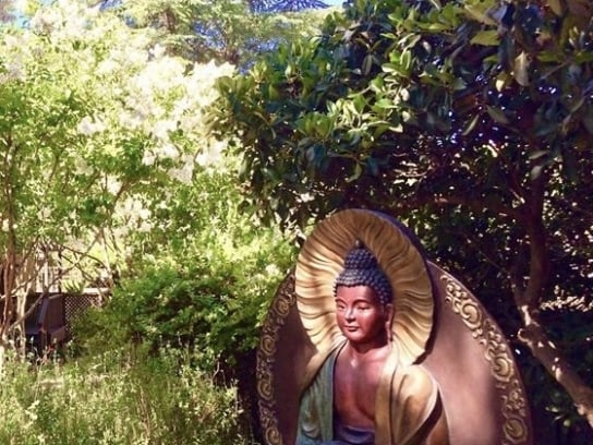A colorful Amitabha Buddha statue sitting in meditation within a lush, sunlit garden setting.