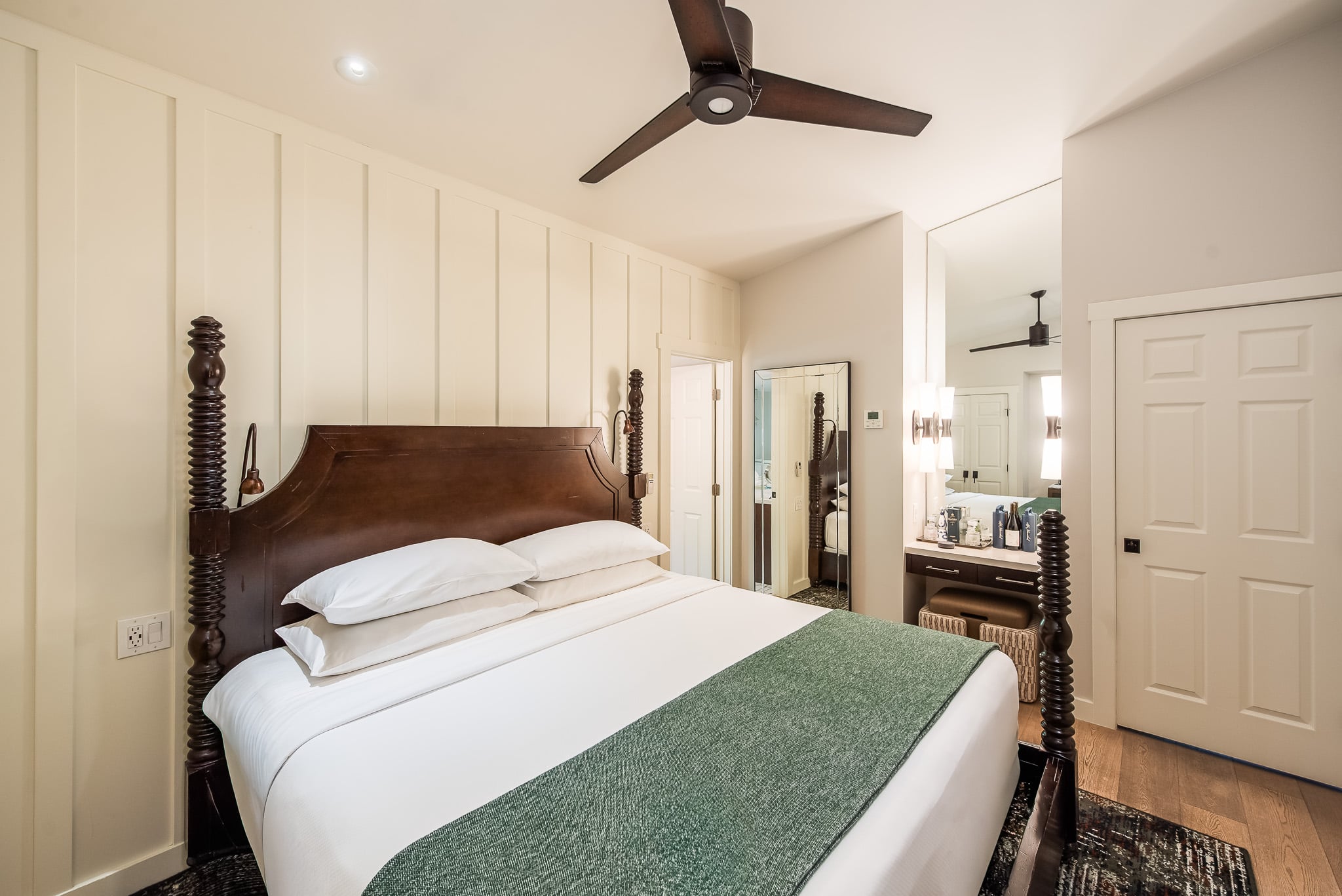 A hotel room with a bed, ceiling fan, door, mirror, and vanity.