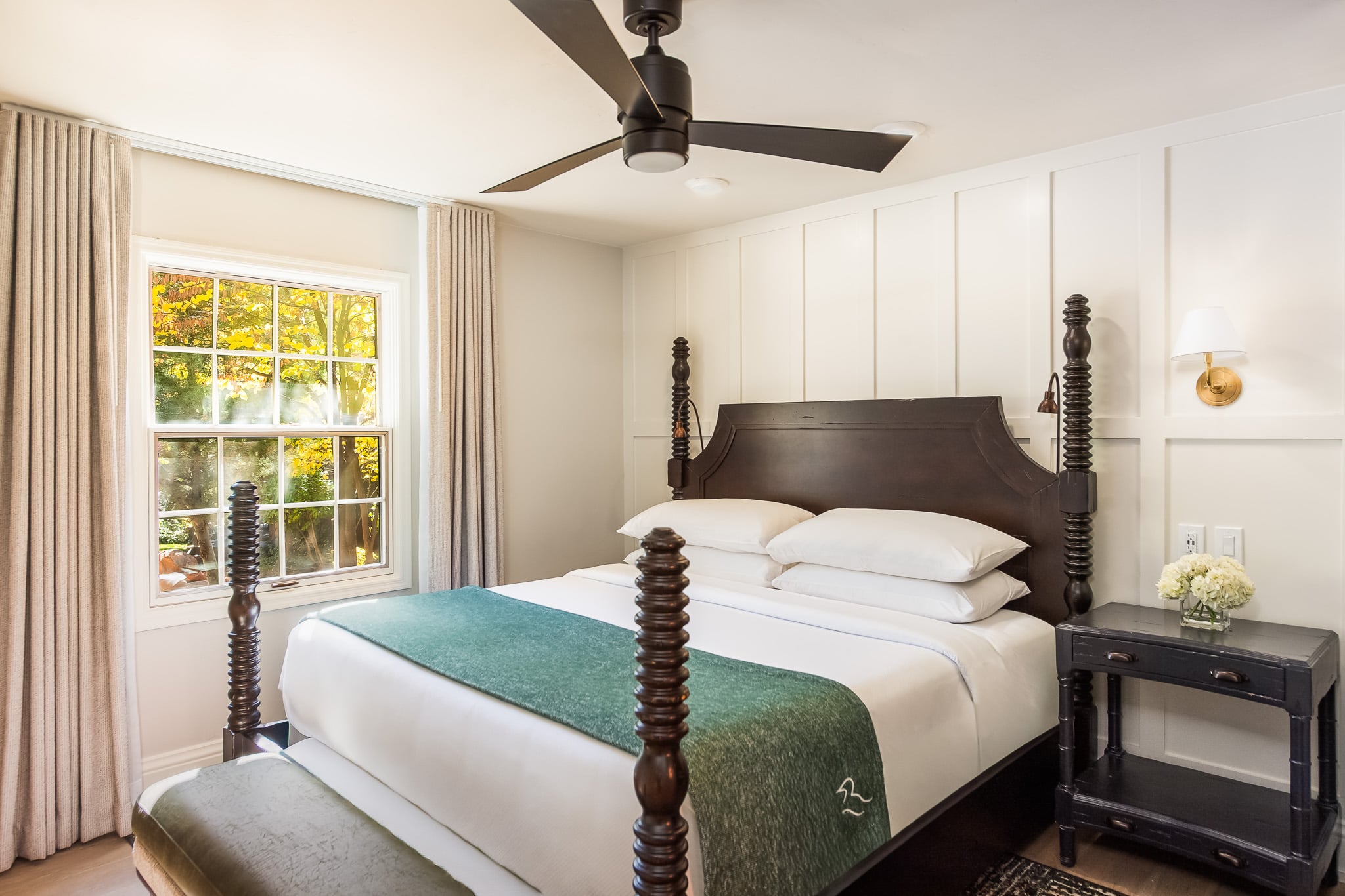 A wide view of a hotel room with a large bed, nightstand, and window showing a view of trees outside.