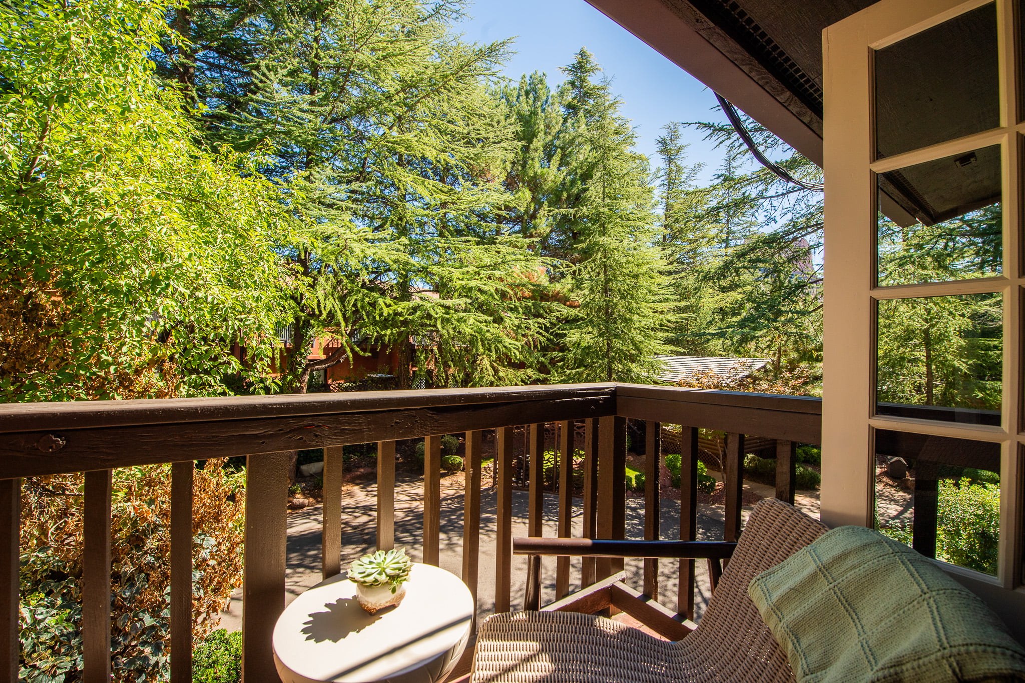 A balcony with a wicker chair, a green plaid pillow, and a small round table with a succulent plant, overlooking trees and a clear blue sky.