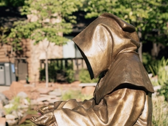 A close-up of a golden, hooded statue of St. Francis meditating by a tranquil garden pond.