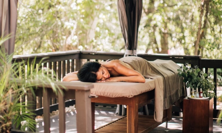 A woman lies on a massage table outdoors, surrounded by plants.