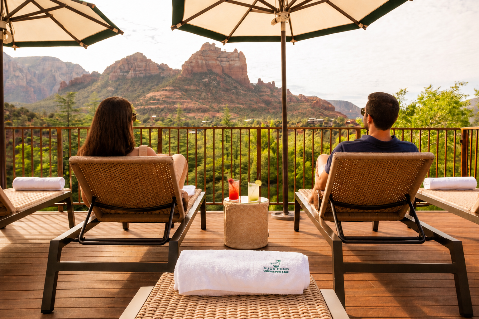 Couple lounging on wicker chairs on a wooden deck under large umbrellas, mountains in the distance.