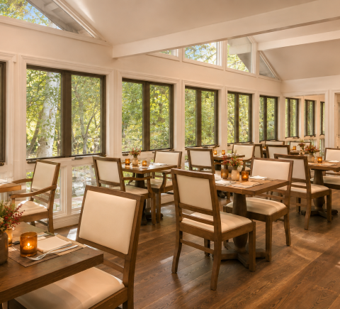 Cozy sunlit restaurant with large windows, wooden tables and cream upholstered chairs, candles on tables.