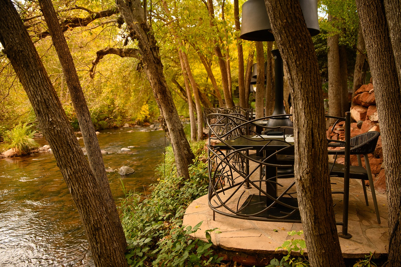 Outdoor riverside seating area with metal high-top chairs around a round table on a stone patio, surrounded by trees and autumn foliage.